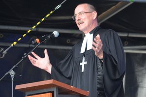 Ein Mann in geistlicher Kleidung mit umgehängtem Kreuz spricht mit ausgebreiteten Armen in ein Mikro