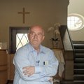Herr Zander steht in einem hellen Hemd in einem Treppenhaus, die Arme vor der Brust verschränkt, im Hintergrund ein Kreuz an der Wand.