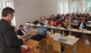 Blick vom Rednerpult auf viele Menschen, die an Tischen in Reihen sitzen. Ein Mann steht am Rednerpult