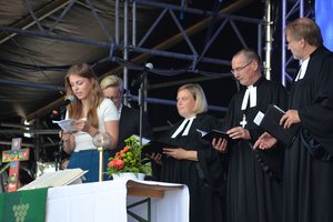 Eine junge Frau mit weißem Shirt spricht in ein Mikro, daneben stehen drei Menschen in geistlicher Kleidung